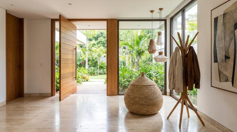 Entrada de casa sofisticada vista por inteiro, com cesto de fibra grande no chão para bolsas e um cabideiro elegante com alguns casacos, ambiente claro e de padrão alto