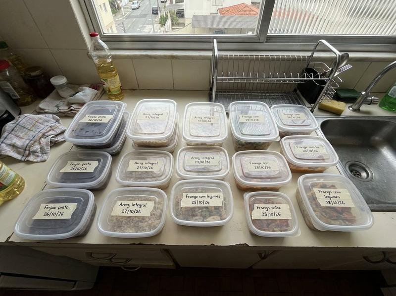 Bancada de cozinha comum vista de cima em ângulo aberto, com marmitas em potes retangulares empilhados e etiquetas de fita crepe escritas à mão com nome do prato e data